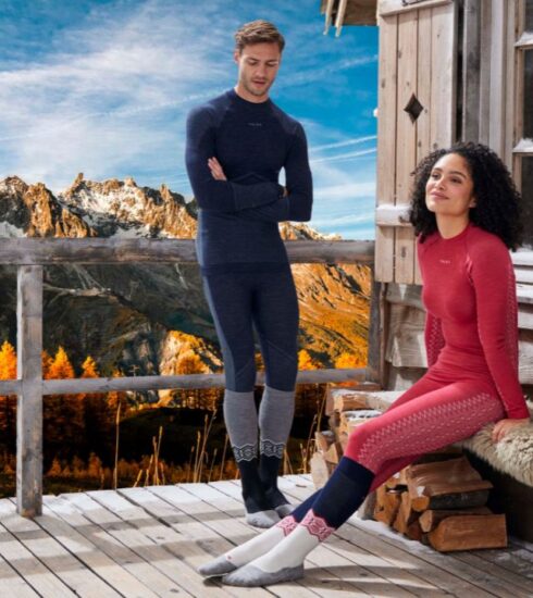 Due persone indossano abbigliamento termico FALKE ESS davanti a un paesaggio montano autunnale, con montagne innevate sullo sfondo e alberi colorati di arancione.