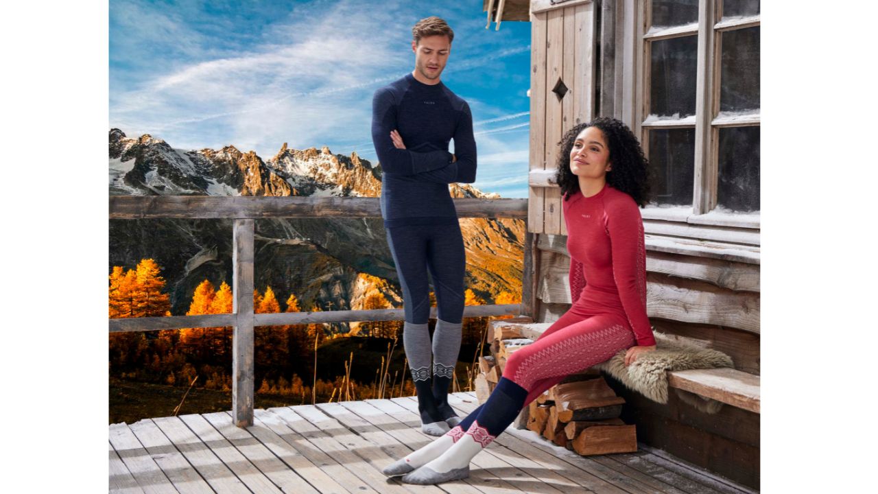 Due persone indossano abbigliamento termico FALKE ESS davanti a un paesaggio montano autunnale, con montagne innevate sullo sfondo e alberi colorati di arancione.