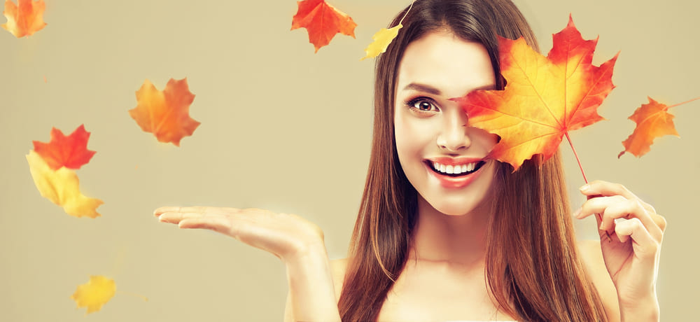 Donna sorridente con foglie autunnali e capelli lucenti, rappresentazione della cura dei **capelli in autunno** per mantenere la chioma sana e splendente.